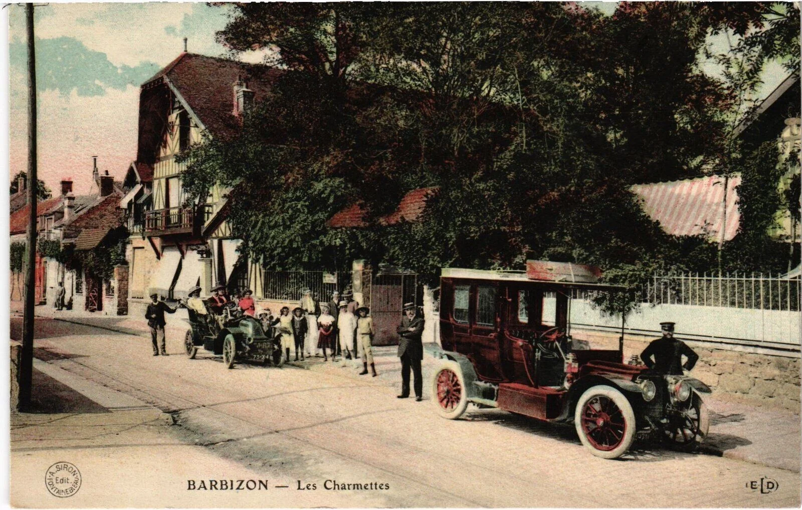 Carte postale colorisée des Charmettes vers 1910 : façade à colombages sur la Grande Rue, deux automobiles d'époque et un groupe d'habitants devant le portail.